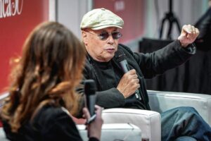 Scrittore con cappello e occhiali da sole parla al microfono durante un incontro letterario, con banner rosso sullo sfondo