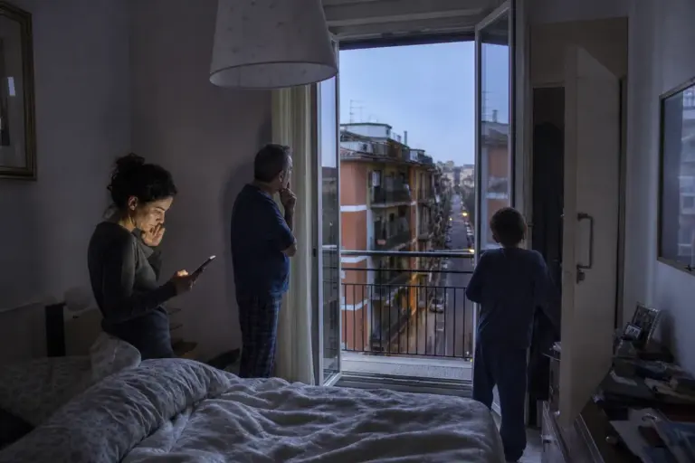 Famiglia in camera da letto all’alba: donna al telefono, uomo e bambino sul balcone dopo una scossa