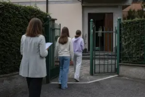 Assistente sociale con cartellina accompagna due ragazze verso l’ingresso di una casa protetta con cancello verde