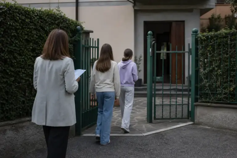 Assistente sociale con cartellina accompagna due ragazze verso l’ingresso di una casa protetta con cancello verde