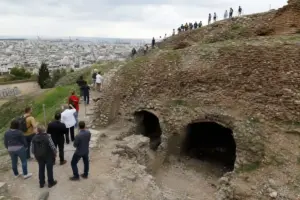 Visitatori in fila su un sentiero che sale una collina di cocci, con grotte alla base e panorama urbano sullo sfondo