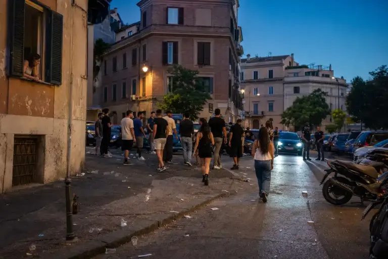Gruppo di ragazzi in strada all’alba tra rifiuti, con un residente alla finestra e polizia sullo sfondo