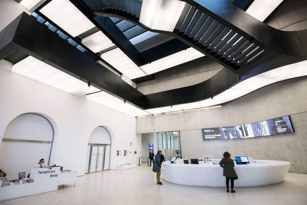 Interno del MAXXI di Roma con la reception curvilinea bianca, visitatori e la struttura architettonica a travi nere incrociate sul soffitto