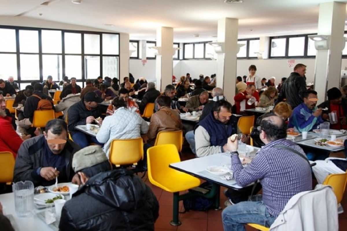 Sala mensa affollata con persone in difficoltà economica sedute ai tavoli durante un pasto assistito