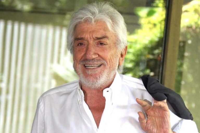 Gigi Proietti sorridente in camicia bianca e giacca sulla spalla