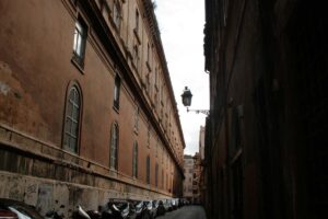 Vicolo stretto del centro storico di Roma con edifici in mattoni rossi, auto parcheggiate e lanterna d'epoca