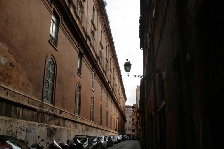 Vicolo stretto del centro storico di Roma con edifici in mattoni rossi, auto parcheggiate e lanterna d'epoca