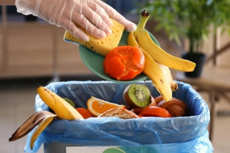 Mano con guanto getta frutta fresca come banane, arance e kiwi in un sacchetto della spazzatura, simbolo dello spreco alimentare