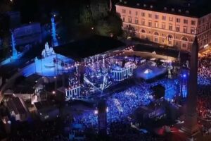 Veduta aerea notturna di Piazza del Popolo a Roma gremita di pubblico durante il concerto del TIM Summer Hits, con palco illuminato di blu