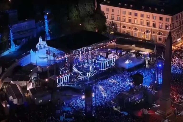 Veduta aerea notturna di Piazza del Popolo a Roma gremita di pubblico durante il concerto del TIM Summer Hits, con palco illuminato di blu
