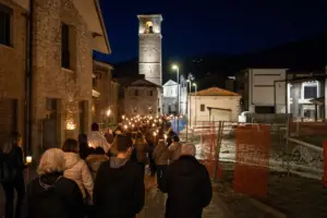 Fiaccolata notturna con cittadini e torce accese lungo una strada del borgo, verso una torre illuminata
