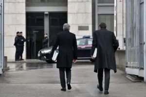 Due avvocati in cappotto scuro con fascicoli camminano verso l’ingresso di un palazzo, con polizia e auto di servizio sullo