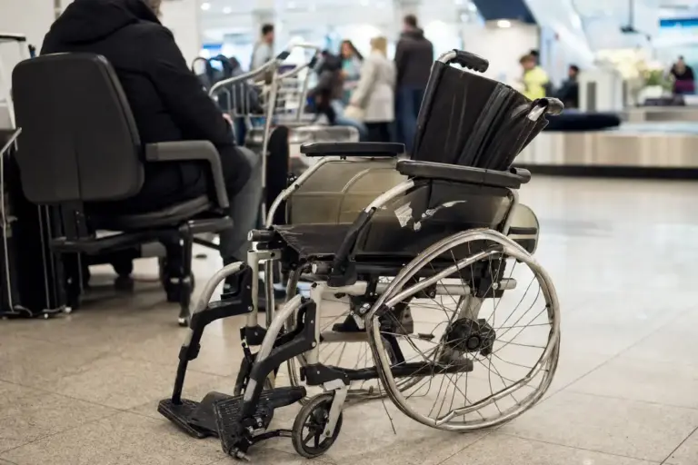 Carrozzina manuale danneggiata nell’area arrivi di un aeroporto, con ruota piegata e passeggeri sfocati sullo sfondo