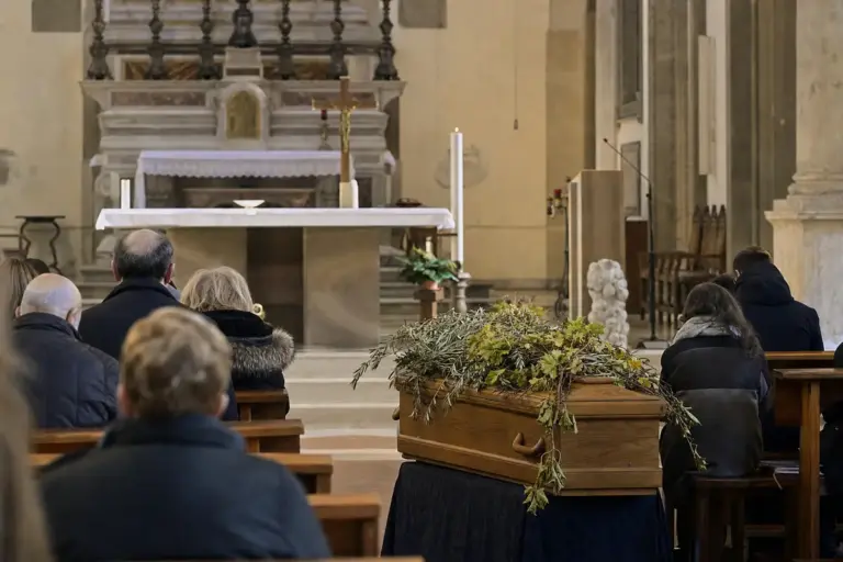 Bara in legno con rami di olivo e tralci di vite in una basilica, con fedeli seduti ai banchi durante il funerale