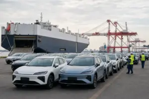 File di auto nuove in un terminal portuale davanti a una nave cargo, con operatori in giubbotti catarifrangenti