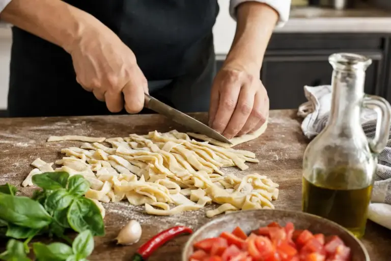 Mani che tagliano striscioline di pasta fresca su tagliere infarinato, con basilico, pomodori, aglio e olio