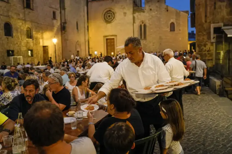 Volontario in camicia bianca serve piatti ai tavoli di una cena comunitaria in una piazza storica illuminata