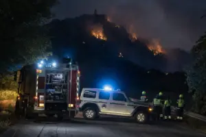 Camion dei Vigili del Fuoco e mezzo di Protezione Civile bloccano una strada di notte mentre un incendio brucia sul monte