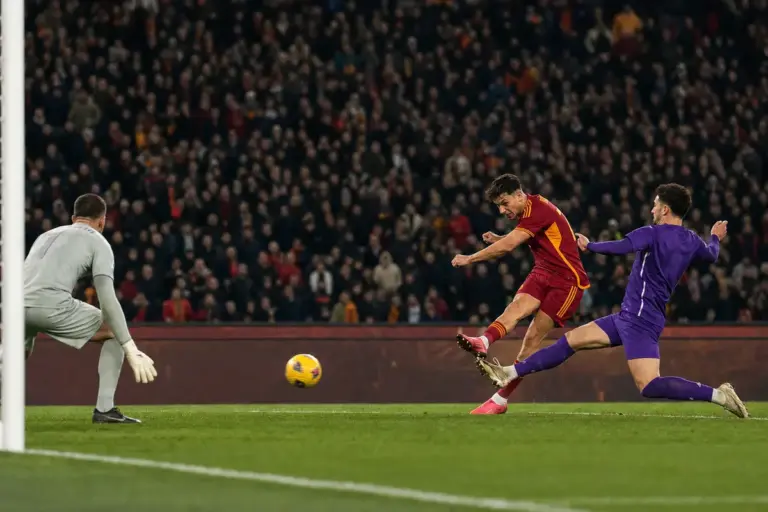 Attaccante in maglia rosso-gialla calcia in area con un difensore in viola in scivolata e il portiere pronto