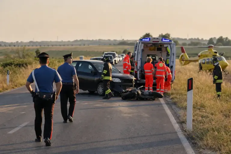 Soccorsi e carabinieri su strada di campagna con auto danneggiata, moto a terra, ambulanza e elicottero sanitario