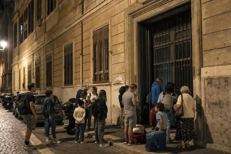 Persone con valigie e borse davanti a un grande portone chiuso di un edificio, di sera, in strada