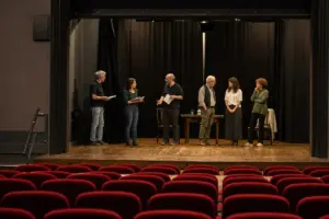 Prove teatrali su un piccolo palco con sei attori e copioni, tavolo e sedie, con poltrone rosse in platea