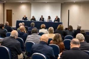 Pubblico seduto ascolta un tavolo di relatori con microfoni in una sala conferenze durante un dibattito politico