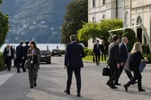 Partecipanti in abito formale davanti a una villa sul lago, con auto scure e delegazioni in movimento