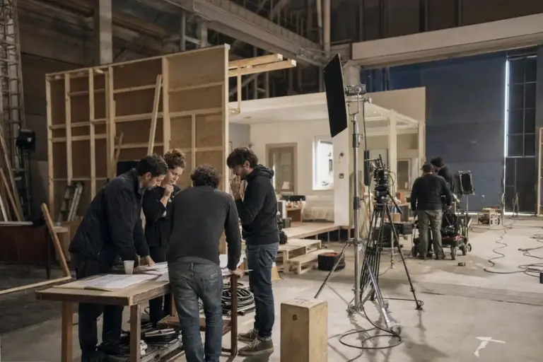 Tecnici su un set in teatro di posa, attorno a un tavolo con planimetrie, con luci, camera su treppiede e scenografia in