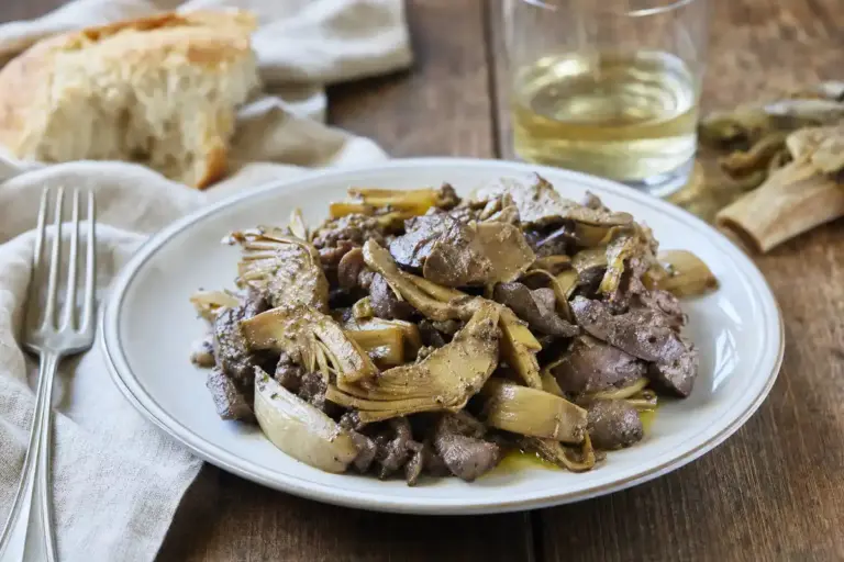 Piatto di coratella d’abbacchio con carciofi su tavolo di legno, con pane rustico, forchetta e bicchiere di vino bianco