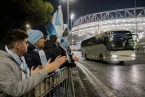 Tifosi con sciarpe biancocelesti applaudono dietro le transenne mentre passa il pullman vicino allo stadio di notte