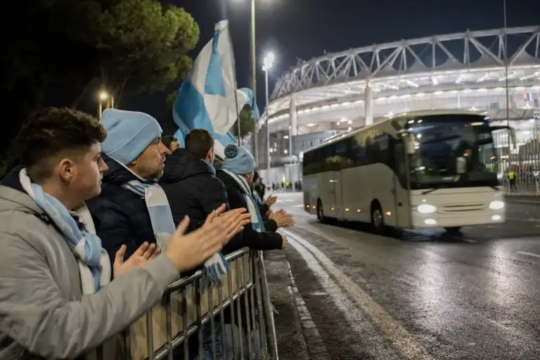 Tifosi con sciarpe biancocelesti applaudono dietro le transenne mentre passa il pullman vicino allo stadio di notte