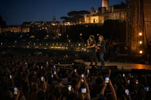 Due musicisti sul palco al tramonto, uno al microfono e uno con chitarra, davanti a una folla con smartphone a Roma