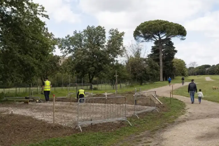 Cantiere con transenne e operai in giubbotti gialli accanto a un sentiero nel parco, con passanti e ciclista