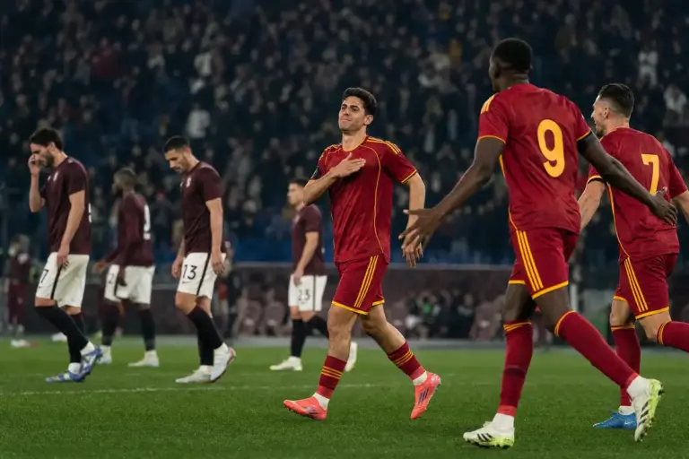 Calciatore in maglia rosso scuro con dettagli gialli celebra con la mano sul petto, mentre gli avversari camminano delusi