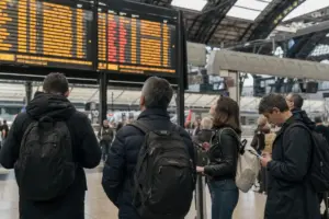 Viaggiatori con zaini controllano il tabellone partenze in stazione, tra possibili cancellazioni e ritardi