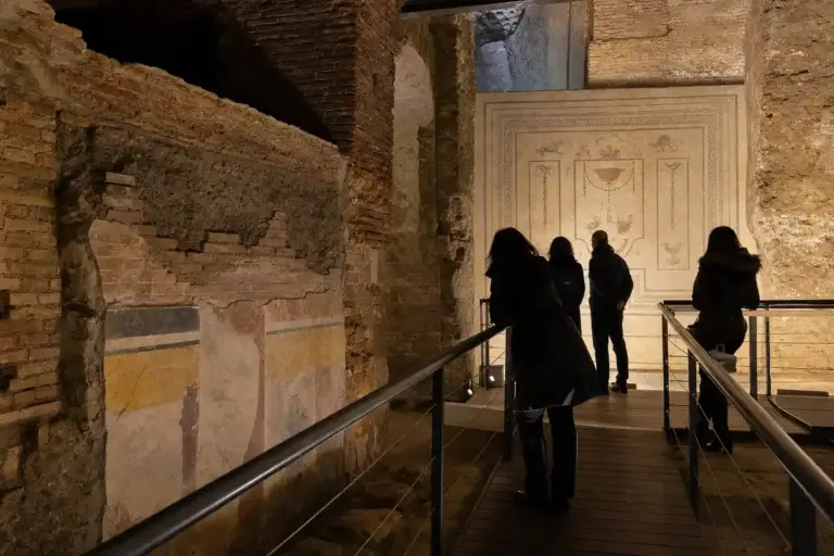 Visitatori su passerella in una galleria archeologica sotterranea con muri in mattoni, affreschi e grande mosaico